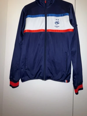 Blå FFF träningsjacka Frankrike - Snygg blå träningsjacka med dragkedja från FFF, Frankrikes fotbollslandslag. Jackan har vita och röda detaljer, broderad tupp och texten 'FRANCE FFF' på bröstet. Tillverkad i glansigt syntetmaterial, perfekta färger för fans av Les Bleus.