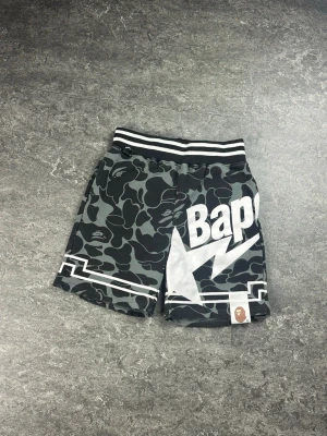 Svarta camo shorts från Bape - Säljer ett par svarta camo shorts från Bape med vit logga och blixtdetalj på ena benet. Shortsen har elastisk midja med svartvita ränder och en liten Bape-etikett framtill. Perfekta för en streetwear-look.