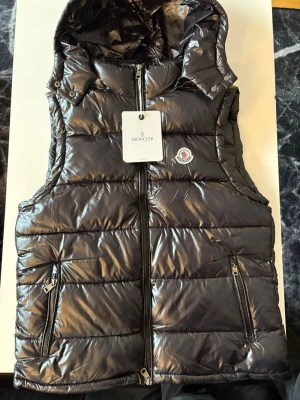 Svart dunväst från Moncler med huva - Snygg svart dunväst från Moncler med glansig finish, två dragkedjefickor och huva. Västen har Moncler-logga på bröstet och stängs med dragkedja framtill. Perfekt för lager på lager och streetwear-vibe.