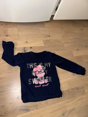 Mörkblå sweatshirt med hundtryck - Mysig mörkblå sweatshirt med tryck av en söt hund med glasögon och texten 'THIS IS MY SWEATER' samt 'woof woof!' i rosa. Tröjan har långa ärmar och ribbade muddar vid ärmslut och nederkant. Perfekt för dig som gillar gullig motiv och avslappnad stil.