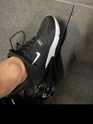 Nike Air Max 270 svarta sneakers - Säljer ett par svarta Nike Air Max 270 sneakers med vit sula och tydlig Air Max-bubbla i hälen. Skorna har mesh-ovandel för bra andningsförmåga, vita detaljer och klassisk Nike Swoosh på sidan. Perfekta för dig som gillar sportig och clean stil.