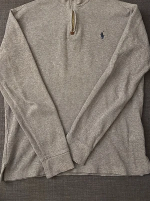 Polo Ralph Lauren 1/4 zip - Säljer en stilren grå långärmad tröja från Polo Ralph Lauren med halvzip och liten blå logga på bröstet. Tröjan har ribbade muddar och är tillverkad i ett mjukt, ribbat material som passar perfekt till vardagslooken.
