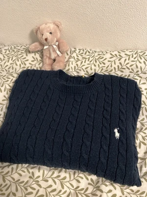 Mörkblå kabelstickad tröja Polo Ralph Lauren - Kabelstickad mörkblå tröja från Polo Ralph Lauren med klassisk rund halsringning och vit broderad logga på bröstet. Tröjan har långa ärmar och ribbade muddar vid ärmslut och nederkant. Perfekt för dig som gillar tidlös och stilren design.