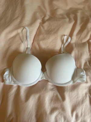 Klassisk Vit bh med bygel - Säljer en enkel och stilren vit bh med justerbara axelband. Bh:n har hak- och hyskknäppning bak och släta kupor utan spets eller mönster, bra under T-shirt! Den har även bygel för extra stöd. Perfekt för dig som vill ha en basic bh med bra stöd också🩷Storlek 70A, aldrig använd så inga fel!! 