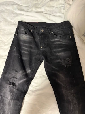 Svarta distressed jeans från Dsquared2 - Säljer ett par svarta jeans från Dsquared2 med slitna detaljer och trasiga partier på benen. Jeansen har fem fickor, knappgylf och en snygg tvättad look. Materialet är klassisk denim och passformen är smal. Perfekta för en trendig och avslappnad stil. Aldrig använt 