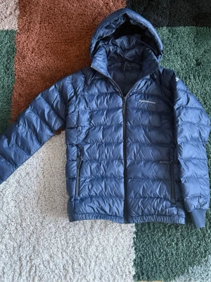 Blå dunjacka från Peak Performance - Snygg blå dunjacka från Peak Performance med huva och dragkedja framtill. Jackan har två sidofickor med dragkedja och quiltad design som håller värmen. Perfekt för kalla vinterdagar och riktigt skön att bära. Klassisk logga på bröstet. Har lagat med en lapp på baksidan men syns ej. Hör av dig om du har några frågor!
