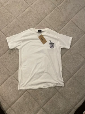 Vit Stüssy t-shirt med tryck - Säljer en vit t-shirt från Stüssy med coolt tryck på bröstet och stort grafiskt motiv på ryggen. T-shirten har korta ärmar och är gjord i mjuk bomull. Perfekt för dig som gillar streetwear och vill sticka ut med en unik design.