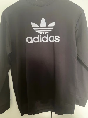 Svart sweatshirt från Adidas - Svart sweatshirt från Adidas med det klassiska vita Trefoil-logotypen och texten på bröstet. Tröjan har rund halsringning och långa ärmar. Perfekt för en sportig och avslappnad stil.