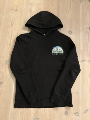  Casablanca hoodie - Säljer en svart hoodie från Casablanca med färgglad grafik och text på både bröstet och ryggen. Trycket visar en byggnad med palmer och blå himmel, samt texten 'CASABLANCA'. Hoodien har huva och magficka, perfekt för en chill och stilren look. Är storlek S men passar M