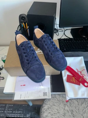 Mörkblåa sneakers från Christian Louboutin - Snygga mörkblåa sneakers i mocka från Christian Louboutin. Skorna har lågt skaft, matchande mörkblå snörning och diskreta detaljer på sidan. Yttersulan är också mörkblå och insidan är ljusbeige. Perfekta för dig som gillar stilrena och exklusiva sneakers.
