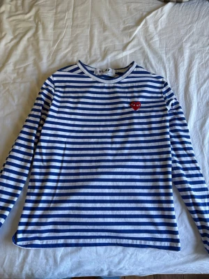 Randig långärmad t-shirt från Comme des Garçons - Säljer en blåvit randig långärmad t-shirt från Comme des Garçons Play med det ikoniska röda hjärtat broderat på bröstet. Tröjan har rund hals och är gjord i mjuk bomull, perfekt för en stilren och avslappnad look.