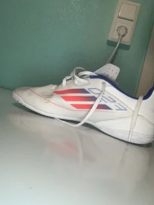 Vita Adidas fotbollsskor med blå och röda detaljer - Säljer ett par vita Adidas fotbollsskor med blå och röda detaljer på sidan och hälen. Skorna har snörning och låg profil, perfekt för snabba rörelser på planen. Tillverkade i syntetmaterial med mönstrad sula för bra grepp.