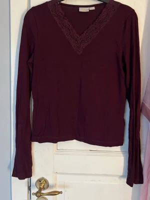Vinröd långärmad topp Gina Tricot - Vinröd långärmad topp från Gina Tricot med v-ringning och spetsdetalj längs halsringningen. Toppens material känns mjukt och stretchigt, perfekt för en snygg och bekväm look. Passar dig som gillar stilrena plagg med en twist.