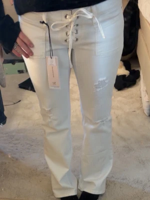 Vita lace-up jeans med slitningar - Säljer ett par vita jeans från 14+ med snörning i midjan och coola slitningar på benen. Modellen har raka ben och fickor framtill. Materialet är denim och jeansen har en trendig, avslappnad vibe. Perfekta för dig som vill sticka ut lite extra! Säljes för dyrare pris pga stor efterfrågan men buda gärna! 