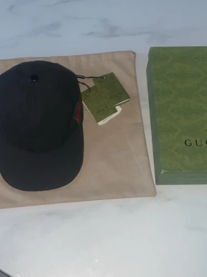 Svart keps från Gucci - Säljer en svart keps från Gucci med klassisk böjd skärm och diskret grön-röd rand på sidan. Kepsen har justerbar rem bak och kommer med originalkartong och tags. Perfekt accessoar för en trendig och stilren look.
