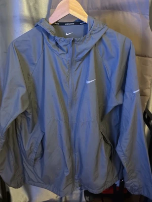 Grå vindjacka från Nike Running - Snygg grå vindjacka från Nike Running med huva och dragkedja framtill. Jackan har två sidofickor och en diskret Nike-logga på bröstet. Tillverkad i lätt och vindtätt material, perfekt för löpning eller utomhusaktiviteter.