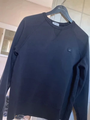 Svart sweatshirt från Acne Studios - Svart sweatshirt från Acne Studios med rund halsringning och det klassiska Face-logotypen på bröstet. Tröjan har en enkel och stilren design med ribbade muddar vid ärmslut och nederkant. Perfekt för en clean och avslappnad look.