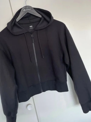 Svart hoodie med dragkedja från Uniqlo - En svart hoodie från Uniqlo med dragkedja framtill och justerbar huva med snören. Modellen är croppad och har breda muddar vid ärmslut och nederkant. Perfekt för en avslappnad och stilren look.