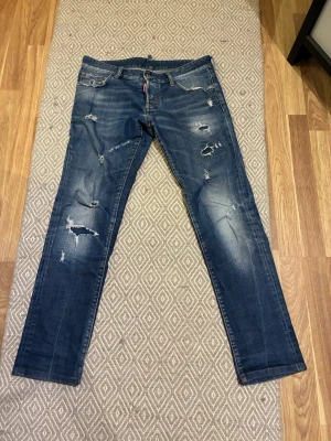 Blå jeans från Dsquared2 - Säljer ett par blå jeans från Dsquared2 med snygga slitningar och trasiga detaljer framtill och baktill. Jeansen har raka ben, klassisk femficksmodell och läderpatch med logga bak i midjan. Perfekta för en cool och avslappnad stil.