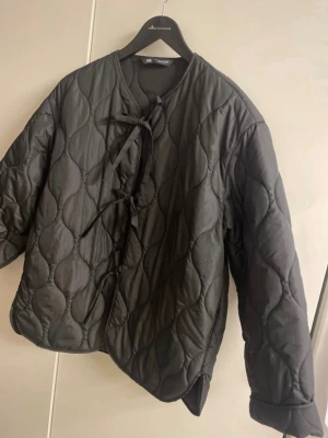 Svart quiltad pufferjacka från Zara - Säljer en svart quiltad pufferjacka från Zara med rund halsringning och knytning framtill. Jackan har ett vågigt mönster i vadderingen och är tillverkad i ett glansigt material. Perfekt för kalla dagar och ger en trendig look.