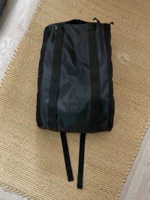 Svart ryggsäck från Db - Säljer en svart ryggsäck från Db med minimalistisk design. Väskan har två breda axelremmar, dragkedja på sidan och är tillverkad i slitstarkt material. Perfekt för resor, träning eller vardagsbruk. 25 Liter.