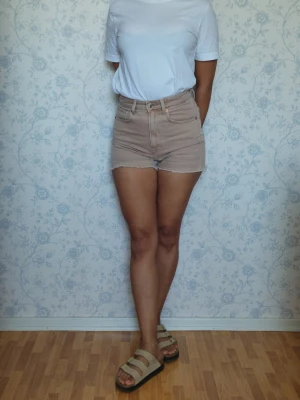 Beige shorts med hög midja  - Säljer ett par beige högmidjade shorts med klassisk femficksdesign och råa benslut. De är tillverkade i bomull och har en normal passform. Perfekta för varma dagar och enkel att matcha med olika toppar.