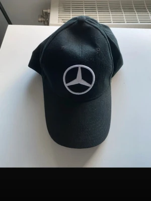 Svart keps med Mercedes-logga - Snygg svart keps med broderad Mercedes-Benz logga framtill. Klassisk böjd skärm och justerbar passform. Tillverkad i bomullstyg som andas. Perfekt accessoar för dig som gillar bilar och streetstyle.