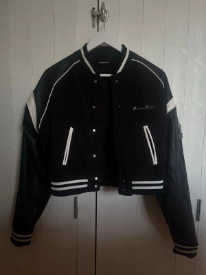 Svart bomberjacka Maniere de Voir - Svart och vit bomberjacka från Maniere de Voir med broderad text på bröstet och en stor patch med Eiffeltornet och en ängel på ryggen. Jackan har vita detaljer på ärmar och muddar samt randiga ribbade avslut. Stängs med tryckknappar och har cool streetwear-vibe. Nypris: 6000