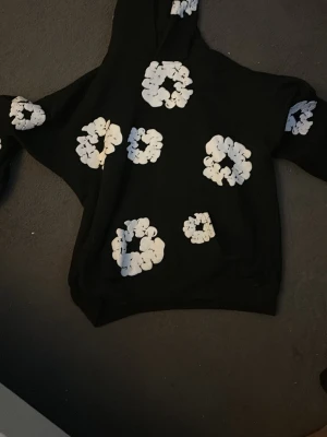Svart hoodie från Denim Tears  - Svart hoodie från Denim Tears med vita blommönster tryckta över hela tröjan, inklusive huvan och ärmarna. Hoodien har en klassisk passform med huva och ribbade muddar. Perfekt för dig som gillar streetwear och unika prints.