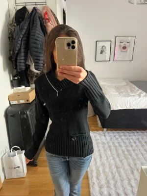Svart stickad tröja med dragkedja - Svart stickad tröja med ribbade muddar och v-ringning med snedställd dragkedja vid halsen. Tröjan har en detalj med liten metallring vid midjan och är långärmad. .