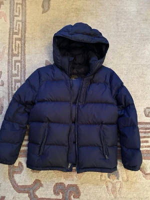  Polo Ralph Lauren - Säljer en mörkblå pufferjacka från Polo Ralph Lauren med huva och flera dragkedjeförsedda fickor. Jackan har broderad logga på bröstet och är perfekt för kalla dagar. 14-16 år ska den passa men jag skulle säga att den passar mellan xs och xxs