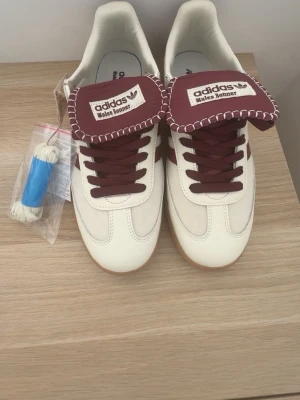 Adidas Wales Bonner Samba sneakers - Snygga Adidas Samba Wales Bonner sneakers i vitt och vinrött med klassiska tre ränder på sidan. Skorna har en unik tunga med kontrastsöm och branding, samt gummisula. Materialmix av läder och mocka ger en exklusiv känsla. Perfekta för dig som gillar streetwear och stilrena detaljer.