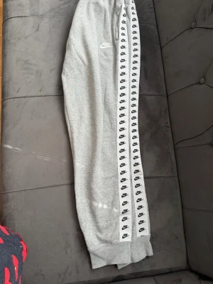 Grå mjukisbyxor från Nike - Snygga grå mjukisbyxor från Nike med vit sidopanel och svarta Nike-loggor längs hela benet. Byxorna har resår i midjan och muddar vid bensluten. Perfekta för en avslappnad och sportig stil.
