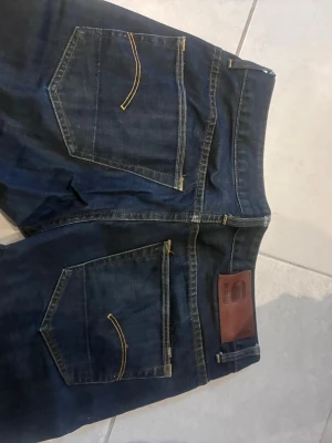 Mörkblå jeans från G-Star RAW - Snygga mörkblå jeans från G-Star RAW med klassisk femficksdesign och kontrasterande gula sömmar. Jeansen har en rak passform och läderpatch med logga bak i midjan. Perfekta för en avslappnad och trendig look.