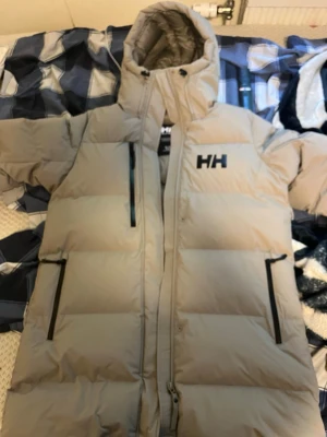 Beige lång dunjacka från Helly Hansen - Säljer en beige lång dunjacka från Helly Hansen med huva och svarta detaljer på dragkedjorna. Jackan har ett stilrent HH-logo på bröstet och ryggen, och är quiltad för extra värme. Perfekt för kalla dagar och riktigt snygg att bära.