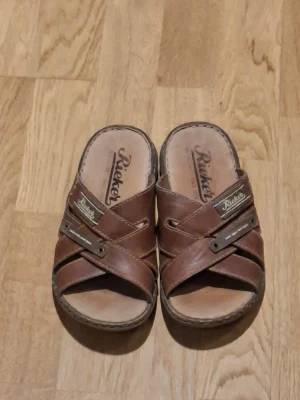 Bruna lädersandaler från Rieker - Säljer ett par bruna sandaler från Rieker med korsade remmar i läder och öppna tår. Sandalerna har en robust sula med mönster och detaljer i metall på remmarna. Perfekta för dig som vill ha en avslappnad och stilren look. De är som nya också men litte använda.