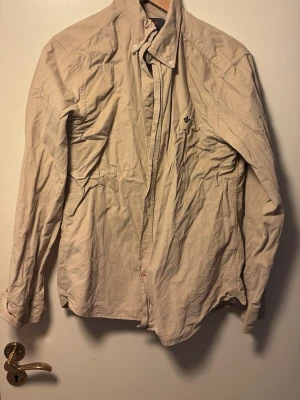 Morris skjorta  - Stilren beige skjorta i manchester med klassisk krage och knäppning framtill. Skjortan har långa ärmar och en liten broderad detalj på bröstet. Perfekt för dig som gillar en avslappnad men ändå snygg look.