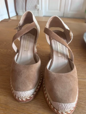 Beige espadriller från Paloma Barceló - Snygga beige espadriller med kilklack och flätad sula från Paloma Barceló. Skorna har en ovandel i mocka och justerbart spänne vid ankeln. Perfekta för en trendig och avslappnad look.