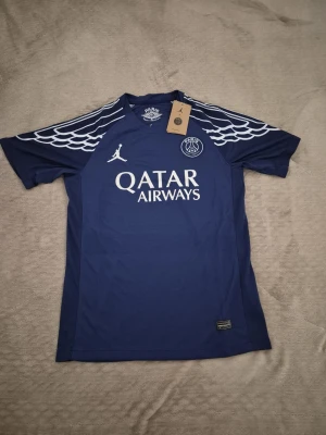 PSG x Jordan blå matchtröja - Säljer en blå Paris Saint-Germain matchtröja från Jordan med vita detaljer och vingmönster på axlarna. Tröjan har tryckt PSG-logga, Jordan-logga och Qatar Airways som sponsor framtill. Perfekt för fotbollsträning eller att bära som supporter. !Kan passa M!