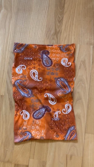 Orange paisley tubtopp  - Säljer en orange tubtopp med paisleymönster i vitt och lila. Toppen är axelbandslös och har en mjuk, stretchig känsla som gör den bekväm att bära. Perfekt för dig som vill sticka ut med färg och mönster. Man kan även ha den som kjol.