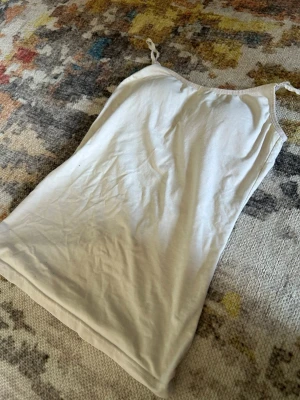 Vit linnetopp med smala axelband - Enkel och stilren beige linnetopp med smala axelband. Toppen är ärmlös och har en rak passform, perfekt att matcha med jeans eller kjol. Tillverkad i mjuk bomull för en bekväm känsla hela dagen.