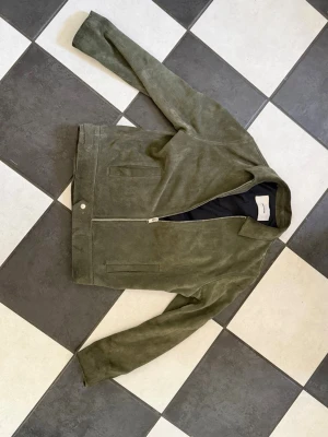 Zadig Voltaire jacka i mocka - Snygg olivgrön skinnjacka i mocka med dragkedja framtill och två fickor. Jackan har klassisk krage och justerbara spännen bak i midjan för perfekt passform. Den är i mocka och är väldigt tung vilket ger en dyr känsla. Perfekt för dig som vill ha en unik jacka till våren. Köpte på Vestiaire för 6500 kr.