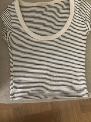 Randig topp från Zara  - Snygg vit t-shirt med tunna svarta ränder och rundad, bred krage. Klassisk och enkel design som passar till det mesta. Tillverkad i mjuk bomull för en skön känsla. Perfekt för dig som gillar stilrena basplagg.