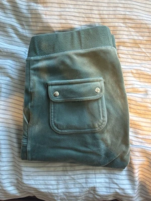 Gröna velourbyxor Juicy Couture - Mysiga gröna mjukisbyxor i velour från Juicy Couture. Byxorna har bred resår i midjan med dragsko, två stora fickor fram och en ficka bak med lock och tryckknappar. Liten Juicy Couture-logga broderad på ena fickan fram.