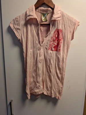 Rosa kortärmad blus med tryck Junk Yard - Ljusrosa blus med korta ärmar och krage från JunkYard.Tryck med röd illustration och stjärnor på bröstet. Blusen har knappar framtill och är tillverkad i ett tunt, skrynkligt bomullstyg. Perfekt för en avslappnad och cool stil.