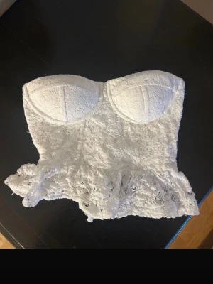 Vit spetsig korsettopp med volang - Superfin vit korsettopp i spets med vadderade kupor och volang nertill. Axelbandslös modell som ger en romantisk och trendig look, perfekt att styla med jeans eller kjol.