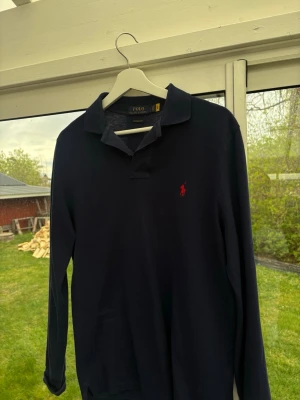 Mörkblå långärmad piké från Polo Ralph Lauren - Klassisk mörkblå långärmad pikétröja från Polo Ralph Lauren med röd broderad logga på bröstet. Skjortan har krage och knappar vid halsen, perfekt för en stilren look.