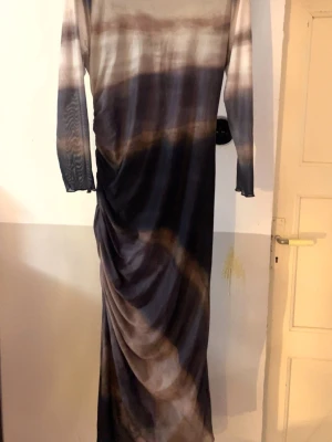Långklänning Shein Curve i mörka toner - Snygg långklänning från Shein Curve i mörka och ljusa toner med ett unikt mönster. Klänningen har lång ärm och draperad detalj på sidan som ger en cool siluett. Perfekt för dig som vill sticka ut med en trendig look.