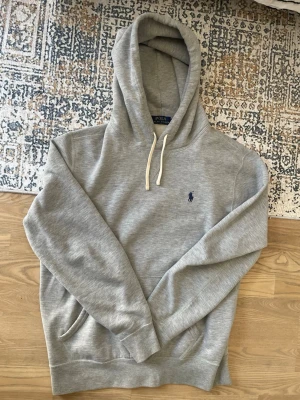 Grå hoodie från Polo Ralph Lauren - Snygg grå hoodie från Polo Ralph Lauren med klassisk blå logga broderad på bröstet. Hoodien har vita snören, känguruficka och ribbade muddar. Tillverkad i mjuk bomullsblandning som känns riktigt skön mot huden. Perfekt för en avslappnad och stilren look.