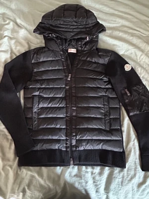 Svart pufferjacka från Moncler - Snygg svart pufferjacka från Moncler med stickade ärmar och huva. Jackan har två dragkedjefickor fram och en Moncler-logga på vänster ärm. Materialmix med quiltad front och rygg i blankt tyg samt ribbstickade muddar och ärmar.